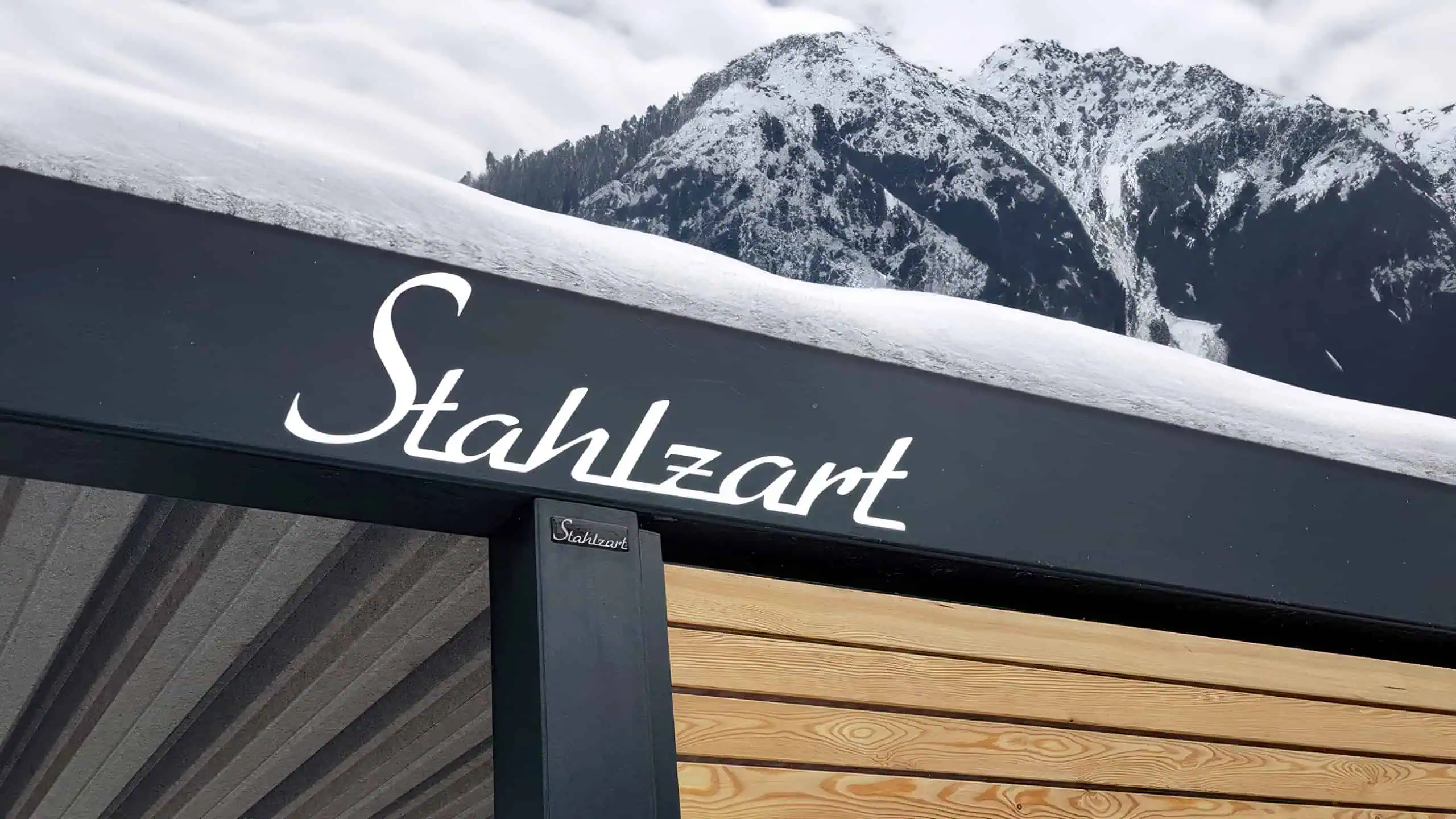 Stahlzart Carport im Winter – Beweis für Stabilität und Langlebigkeit Ein moderner Stahlzart Doppelcarport in Anthrazit mit einer geschlossenen Holzwand, dessen Flachdach sicher eine schwere Schneelast im Winter trägt. Im Hintergrund sind verschneite Berge zu sehen.