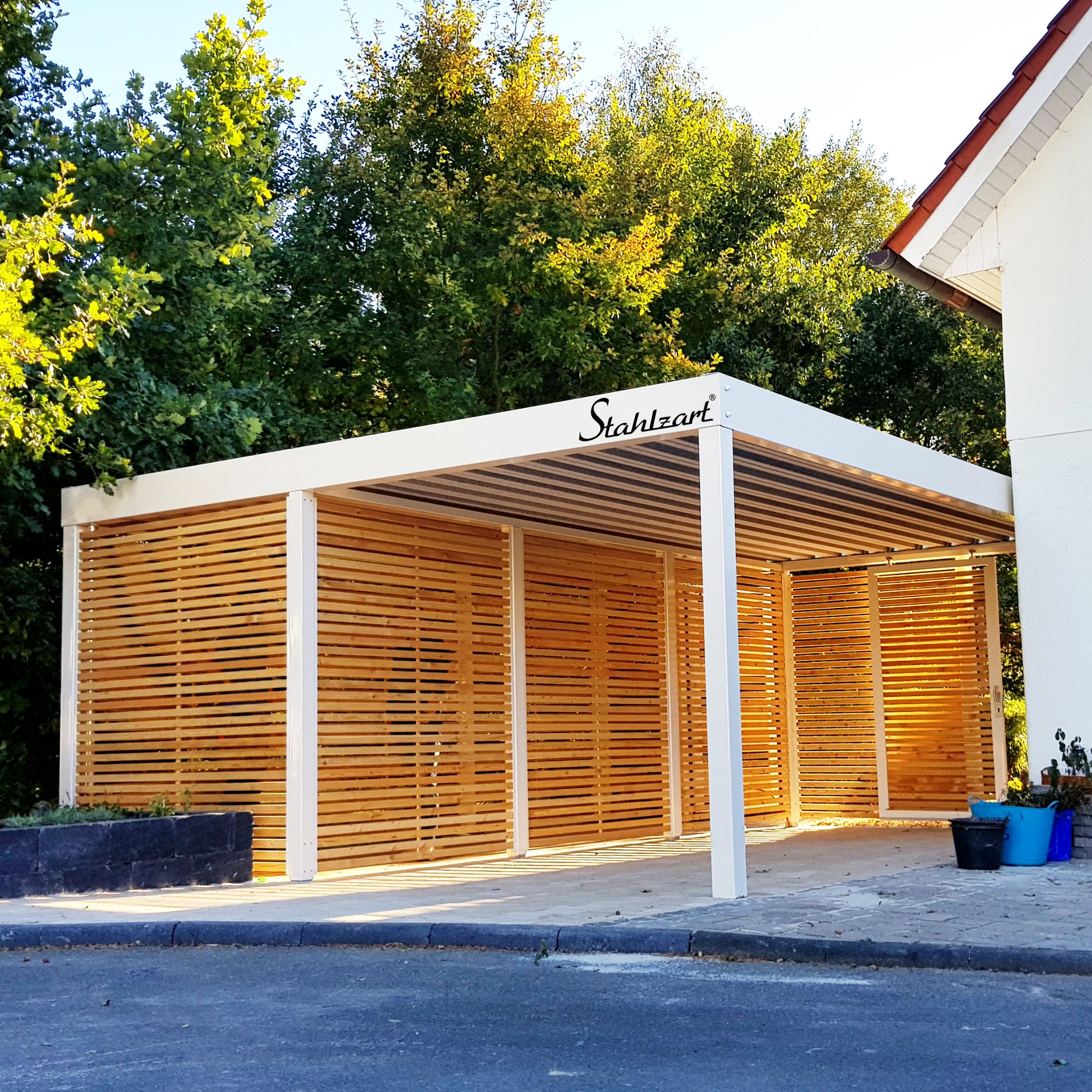 Stahlzart Einzelcarport nach Maß in Weiß Maßgefertigter Einzelcarport von Stahlzart mit einer kompletten Holzverkleidung. Der offene Stellplatz für ein Auto befindet sich rechts, links ist ein voll integrierter Geräteraum. An der Rückseite sind große Schiebetüren als Zugang zu sehen.